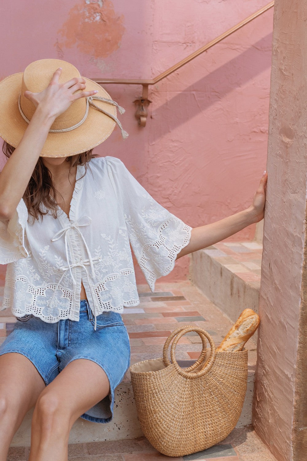 Woman in a white blouse and blue shorts sitting on steps with a pink wall and straw bag.