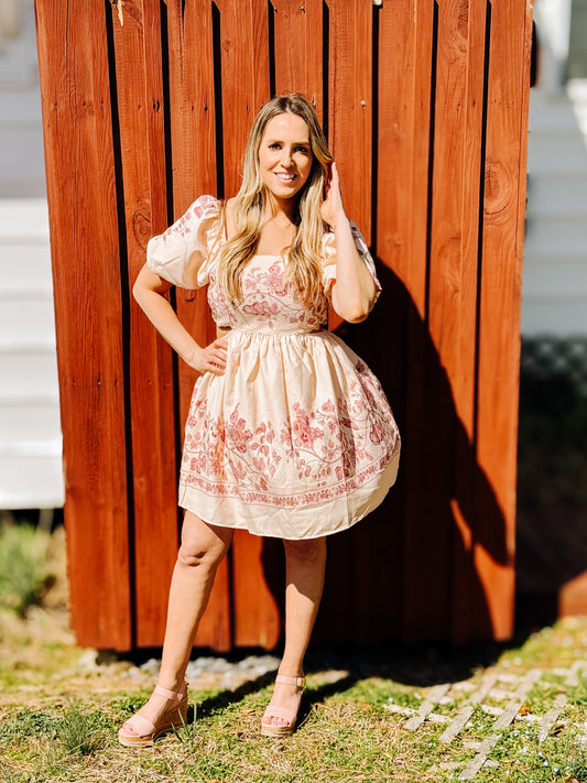 A woman standing outdoors wearing a short, natural color dress with pink floral accents and scallop detailing along the edges.