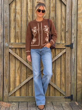 A woman stands in front of a rustic wooden barn door wearing a brown button-front cardigan with cream floral embroidery, light-wash blue jeans, sunglasses, and brown boots, while carrying a dark brown shoulder bag.