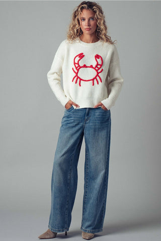 Woman wearing a white sweater with a red crab design and blue jeans on a gray background
