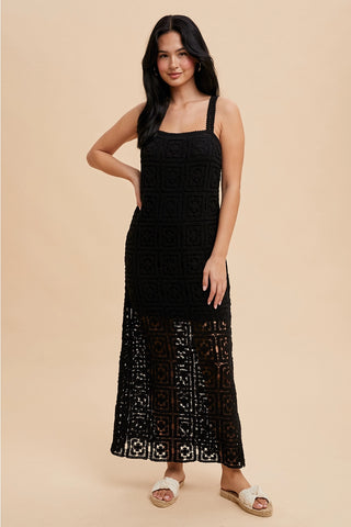 A woman standing in a studio setting, wearing a black square neck maxi dress with tank straps and a concealed side zipper.