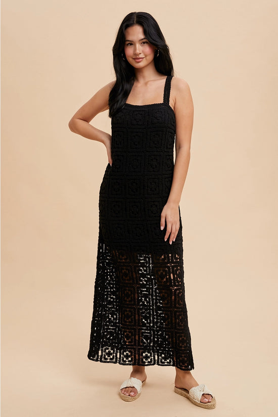 A woman standing in a studio setting, wearing a black square neck maxi dress with tank straps and a concealed side zipper.