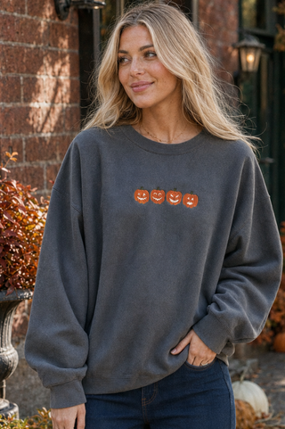 A woman wearing a dark gray crewneck sweatshirt with a row of orange jack-o'-lantern faces across the chest, paired with dark blue jeans, standing outdoors in front of a brick building with pumpkins.