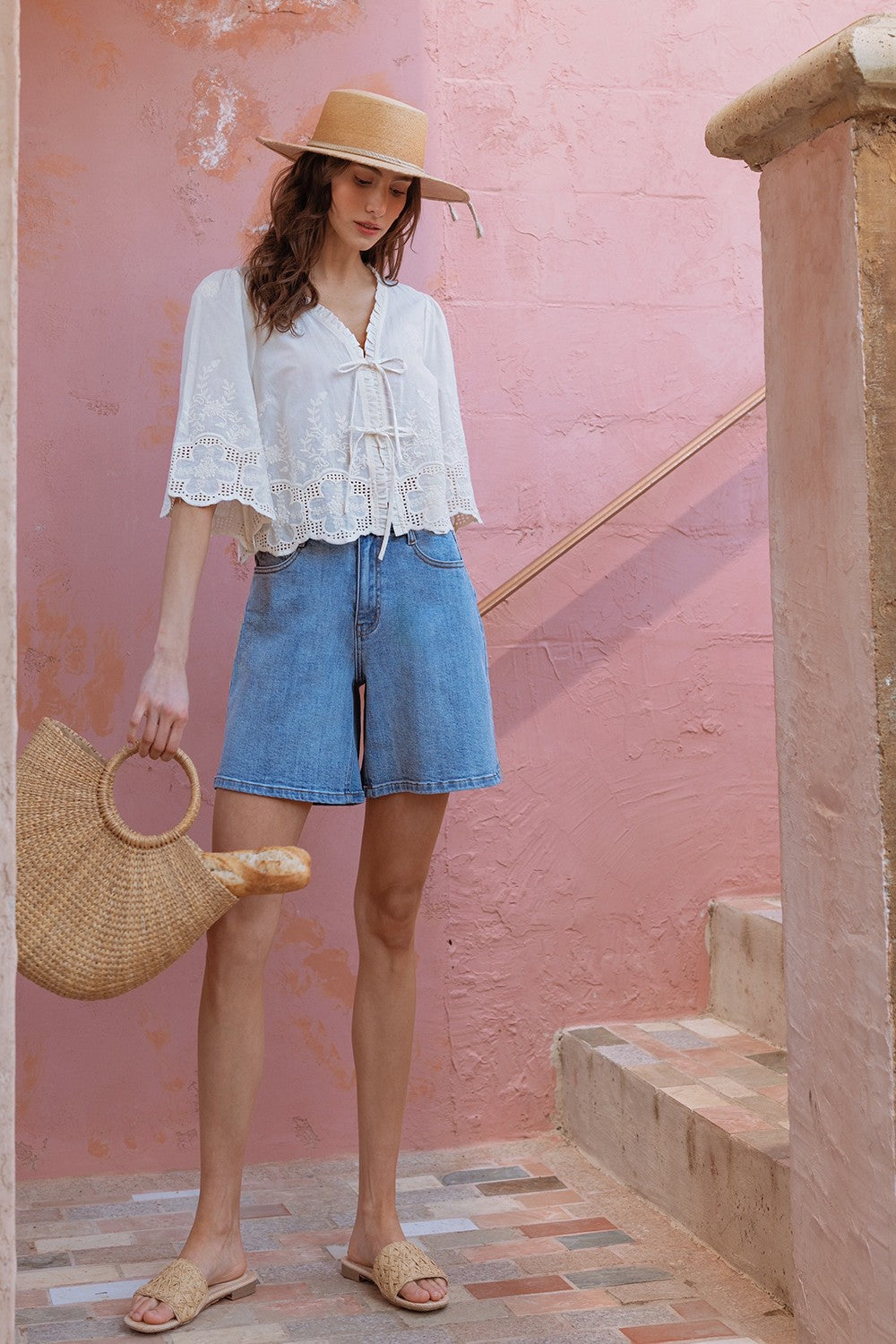 Woman in white blouse and denim shorts standing against a pink wall.