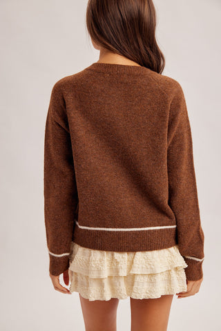 A child wearing a brown knit sweater with white stripe accents at the hem and cuffs, paired with a cream ruffled skirt, shown from the back against a light gray studio background.