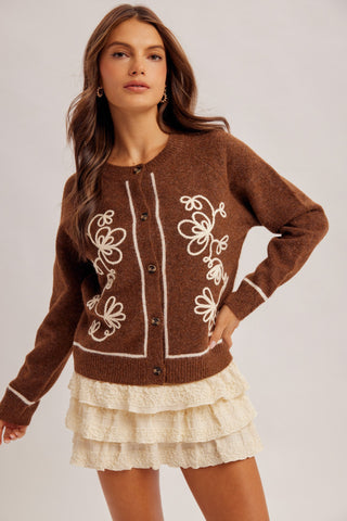 A woman modeling a brown knit cardigan with white floral embroidery and white trim, paired with a cream ruffled skirt, against a light gray studio background.