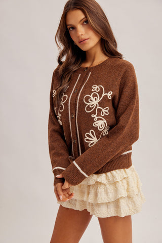 A woman wearing a brown knit cardigan sweater with white floral embroidery and a cream ruffled skirt, photographed against a plain light gray studio background.