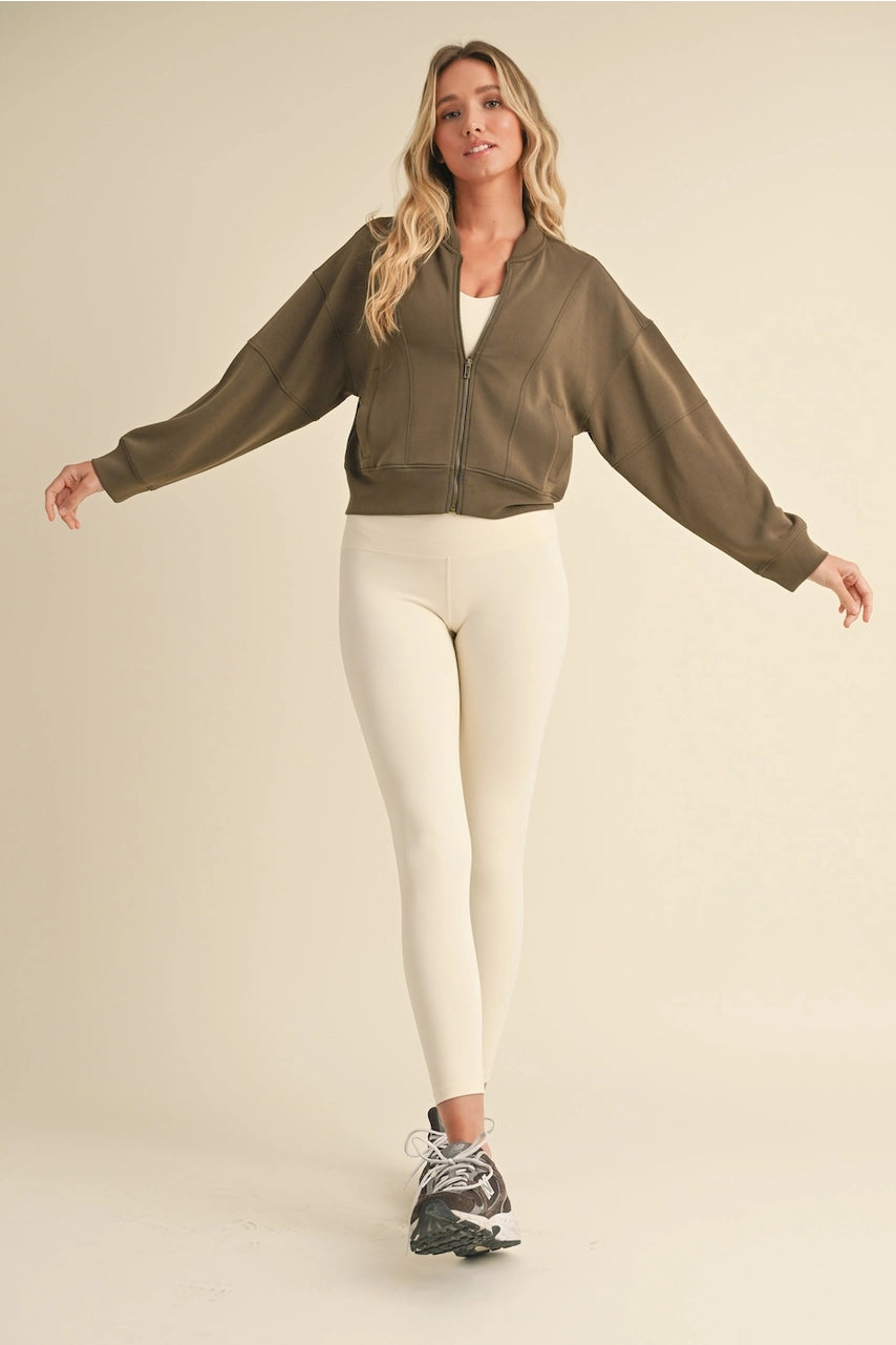 Woman wearing a green bomber jacket and white leggings on a beige background