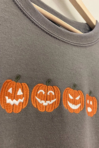A close-up of a dark gray crewneck sweatshirt on a wooden hanger, featuring four embroidered orange jack-o'-lantern pumpkins with white stitched faces across the chest.