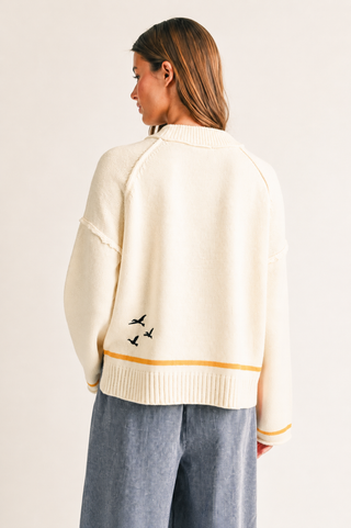 A woman shown from the back wearing a cream knit sweater with a thin mustard yellow horizontal stripe near the hem and small black bird embroidery, paired with blue wide-leg pants, against a plain light background.