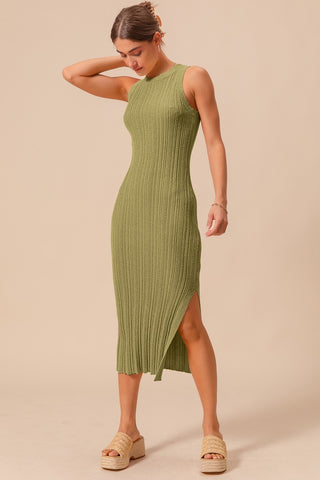 A woman standing wearing an olive green, ribbed, midi dress with a fitted silhouette and a side slit, paired with tan sandals.