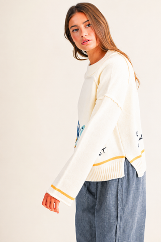 A woman modeling a cream-colored knit sweater with yellow and blue accents, paired with blue jeans, against a plain off-white background.