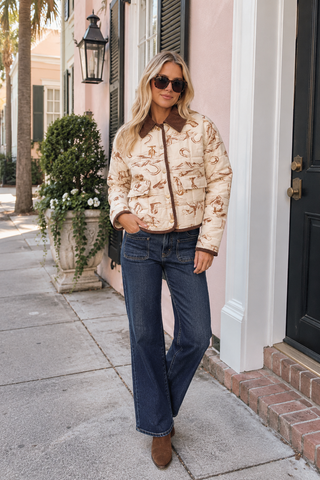 A woman stands on a sidewalk in front of a pink house, wearing a beige bomber jacket with a brown horse-and-carriage print, dark blue jeans, and brown ankle boots.