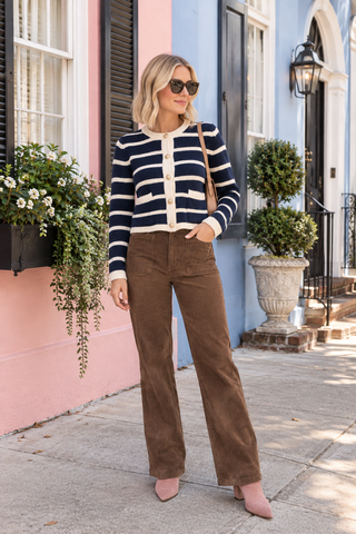 A woman stands on a sidewalk in front of colorful buildings, wearing a navy and cream striped knit cardigan, brown wide-leg corduroy pants, and pink pointed-toe heels, with a tan shoulder bag.