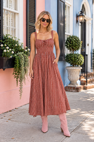 A woman stands on a sidewalk wearing a rust-orange and white plaid midi dress with thin straps and a gathered waist, paired with light pink ankle boots. The background shows colorful building facades in pink and blue with potted plants.