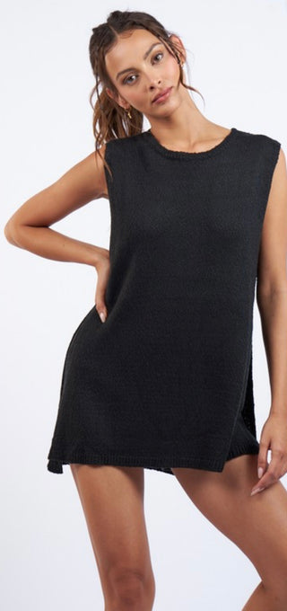 A female model wearing a black knit sleeveless tunic top with an open neckline and paired with elastic waste band shorts.