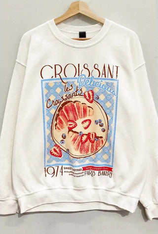 White oversized long-sleeve sweatshirt with a graphic print of a croissant and strawberries on the front, and text that reads 'CROISSANTS LES DELICIA'S'.