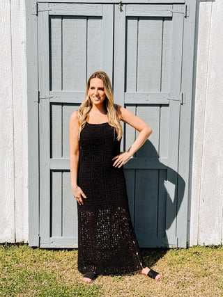 A woman standing in front of a wooden gate, wearing a black square neck maxi dress with tank straps and a concealed side zipper.