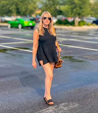 A female model wearing a black knit sleeveless tunic top with an open neckline and paired with elastic waste band shorts.