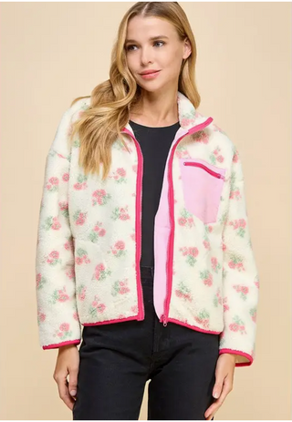 A female model wearing a pink floral print sherpa jacket with a v-neck collar and front zip pocket, paired with black pants.