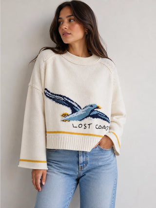 A woman wearing a cream-colored knit sweater with a blue and yellow seagull graphic and the words 'LOST COAST' on the front, paired with light blue jeans, standing against a neutral light gray wall.