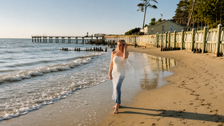 Woman in a white dress walking along a beach with a pier and trees in the background.