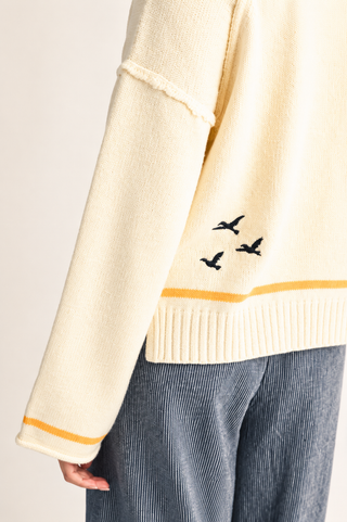 Close-up of a cream knit sweater with three black bird silhouettes embroidered on the lower sleeve, a thin mustard yellow stripe near the hem, and ribbed cuffs and hem, worn with blue corduroy pants.