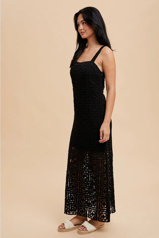 A woman standing in a studio setting, wearing a black square neck maxi dress with tank straps and a concealed side zipper.
