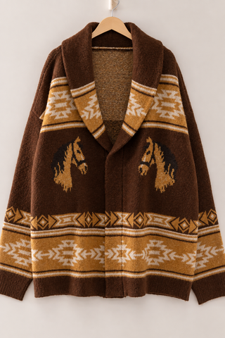 A brown knit cardigan sweater with a shawl collar, featuring geometric patterns and horse motifs in tan and cream, hanging on a wooden hanger against a light gray wall.
